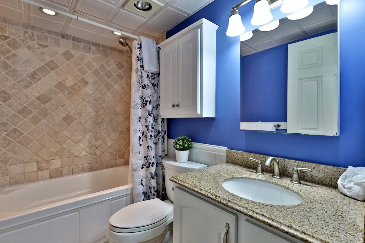 A refreshed, modern bathroom is shown, featuring a bathtub with shower, granite countertop, a well-lit mirror, and a floral shower curtain. The space feels clean, serene, and fully equipped for both relaxing baths and efficient morning routines.