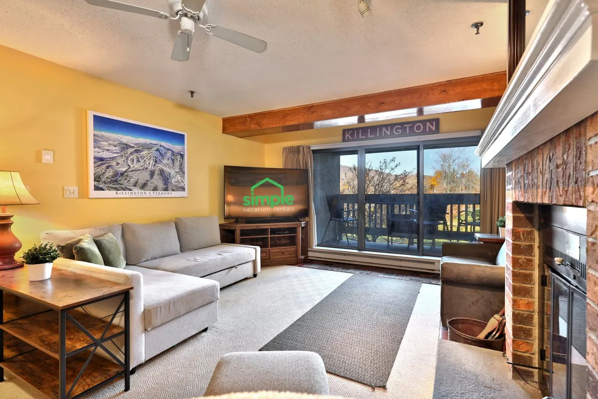 The image showcases a warm and inviting living room with a plush sofa positioned by a crackling fireplace, and a large window framing sweeping mountain views—creating a cozy space perfect for relaxation after outdoor adventures.