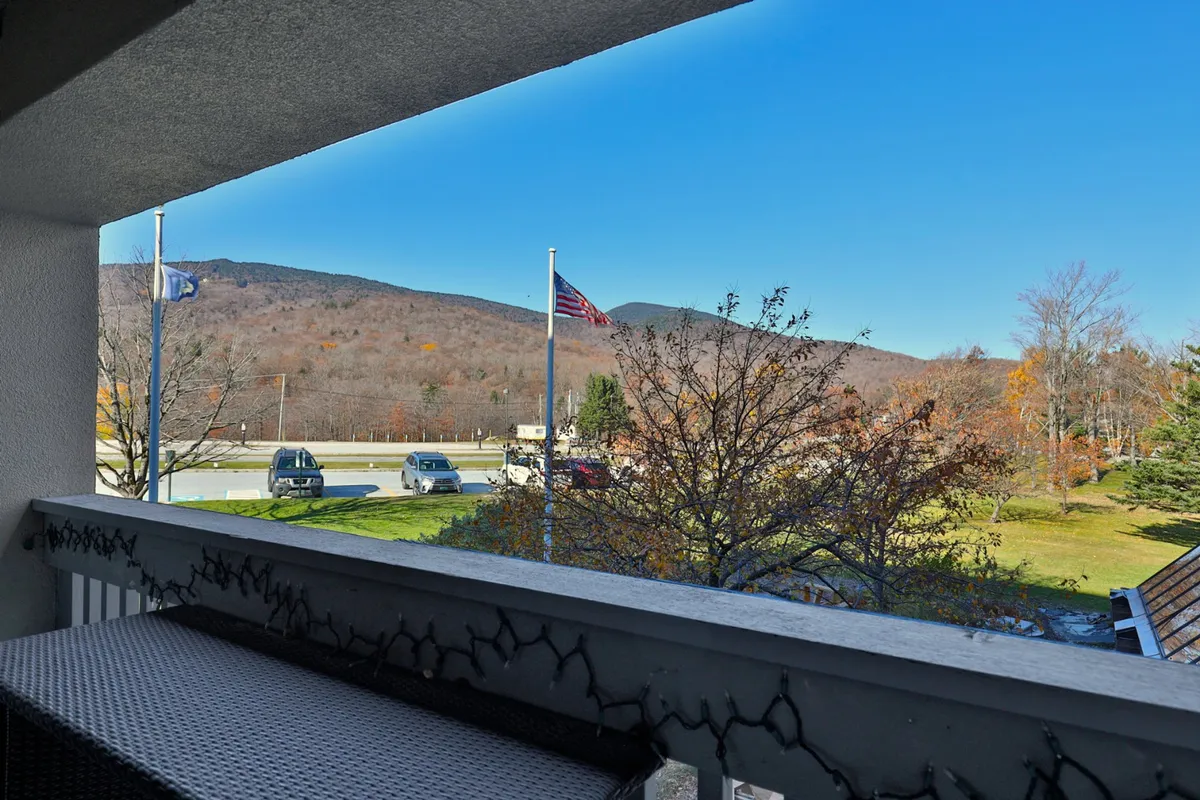 This image captures a panoramic view from a balcony, with a dramatic mountain backdrop, an American flag waving, and a hint of autumn foliage. The balcony railing frames the view, inviting guests to take in the expansive outdoors, watch local events, or simply relax with a book as the day winds down.