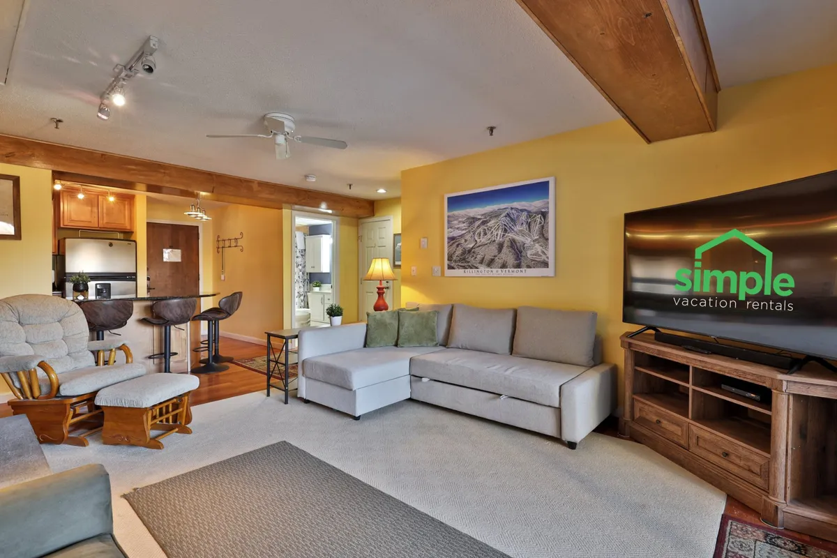 A comfortable living space featuring a soft sofa, a smart TV mounted on the wall, artistic wall décor, and a bar area, all illuminated by natural light. The arrangement encourages both entertainment and relaxation, perfect for streaming shows or spending time with loved ones.