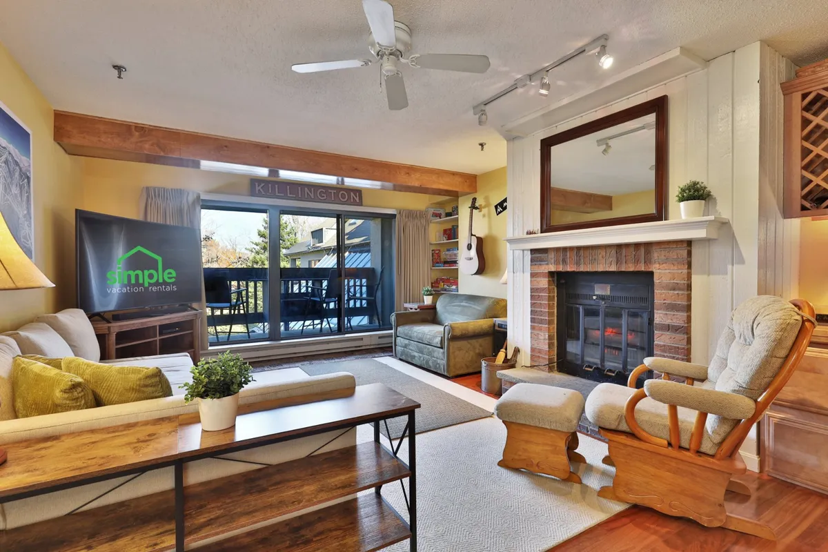 A cozy living room with a welcoming fireplace, a plush sofa, a large window offering a scenic view, a wooden coffee table, and a guitar hanging on the wall. The space is bathed in warm lighting and offers ample seating, emphasizing comfort and togetherness.