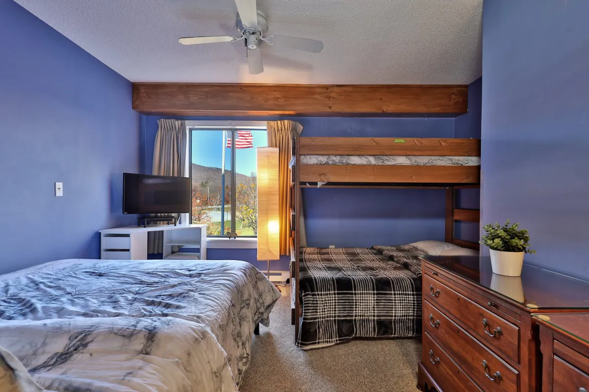 This image shows a spacious bedroom featuring both a full-over-full bunk bed and a plush queen bed, with a large window offering natural light and a view. A wooden dresser and television create a comfortable, family-friendly atmosphere ideal for relaxation. The arrangement is perfect for accommodating families or groups of friends.
