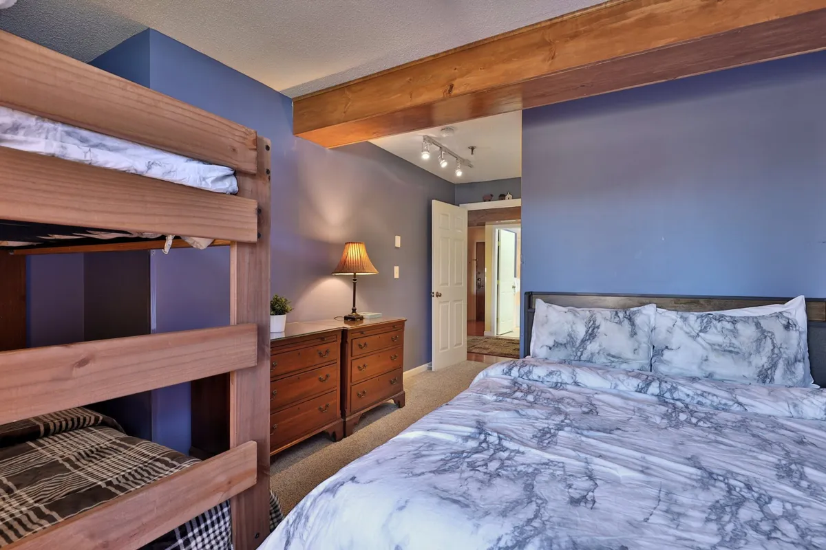 This inviting bedroom features a full-over-full bunk bed alongside a queen bed, complemented by a dresser and bedside lamp. The warm lighting and thoughtful layout create a cozy, welcoming space for shared moments and restful sleep.