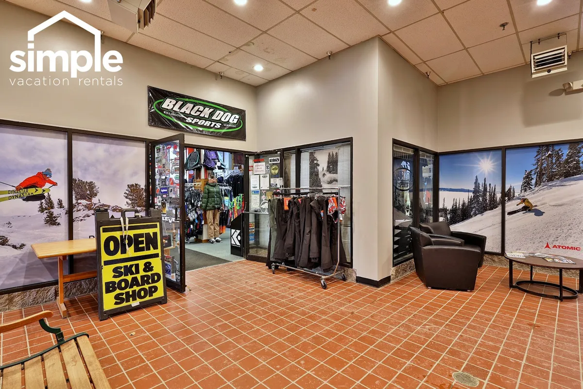 The image shows the entrance to 'BLACK DOG SPORTS,' a ski and board rental shop at the resort. Ski and snowboard gear is displayed prominently, with a seating area and welcoming vibe, ideal for families or groups preparing for their day on the mountain.