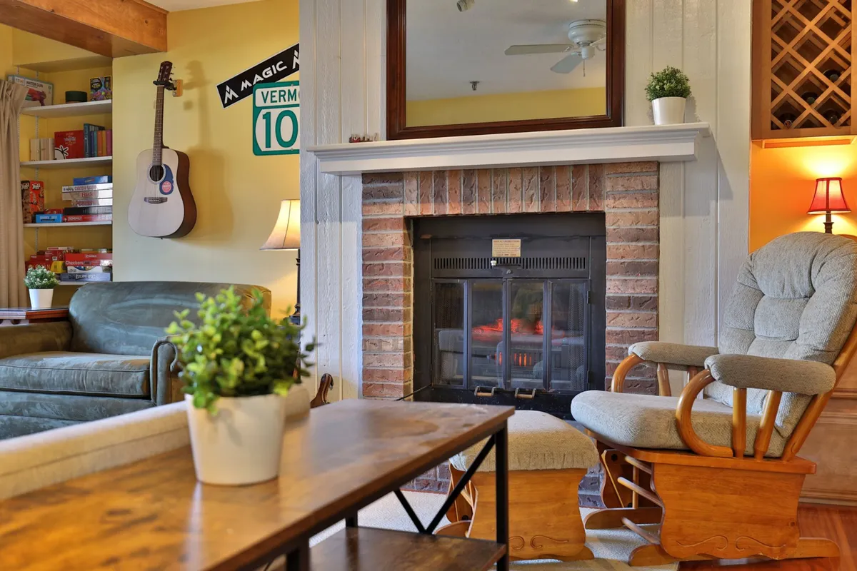 An inviting interior scene featuring a cozy fireplace, a guitar, a rocking chair, and shelves with games. The environment suggests intimate group gatherings, game nights, and relaxing by the fire after a day of adventure.